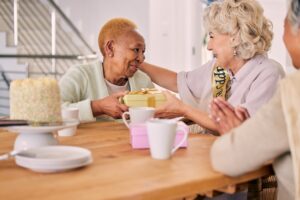Senior friends, birthday cake and women celebration at a home with a present and gift with people.